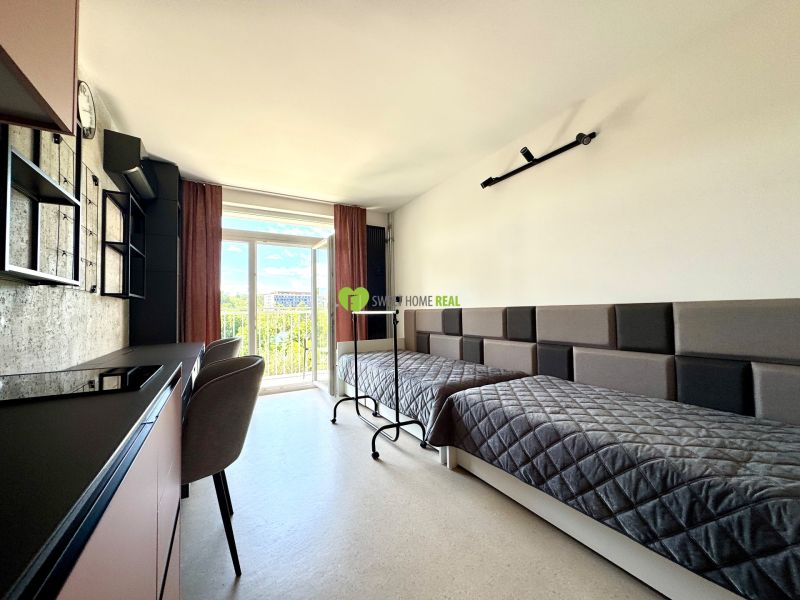 A studio apartment with two beds, a table, and a view through a large window.