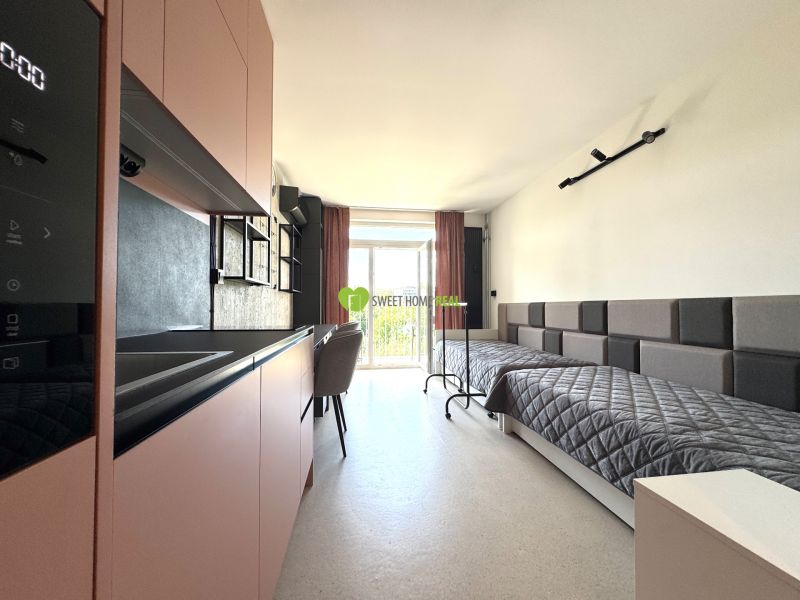 Studio apartment with a bed, kitchenette, and balcony doors, wooden decor flooring.