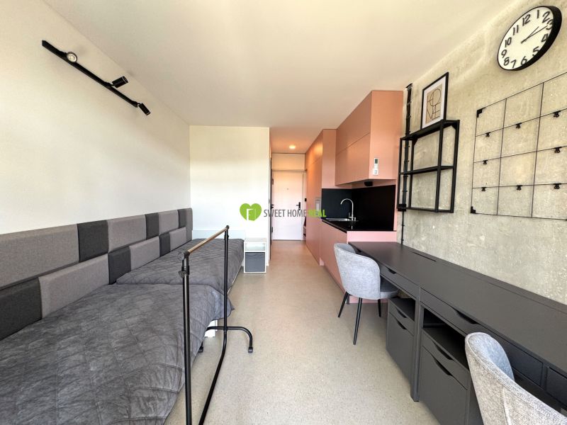 A studio apartment with a bed, pink kitchen, table, and chair in the interior.