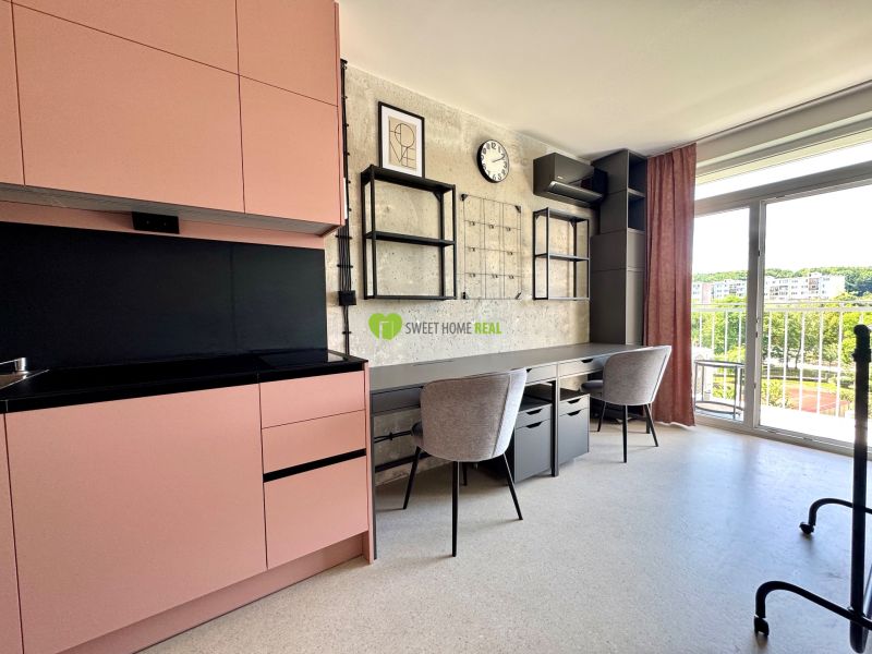 A studio apartment with a pink kitchen unit, a work desk, and a balcony with a view of greenery.