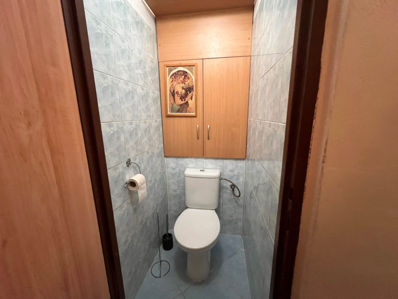 A toilet in a 2-room apartment with blue tiles and wooden decor cabinets.