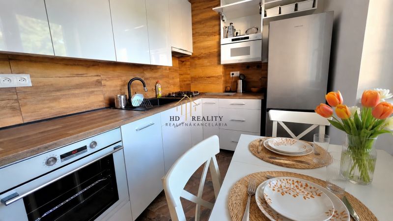 A kitchen in a 2-room apartment with white cabinets, a wood-patterned floor, and a table with flowers.