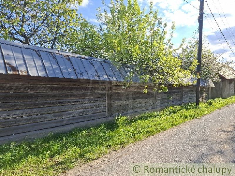 A wooden fence by a family house in Šumiac, surrounded by trees and a road.