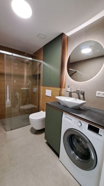 Bathroom of a 2-room apartment with a shower, washing machine, mirror, and cabinets in a neutral color.