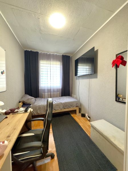 A room in a 4-room apartment with a desk, a bed, and a TV on the wall.