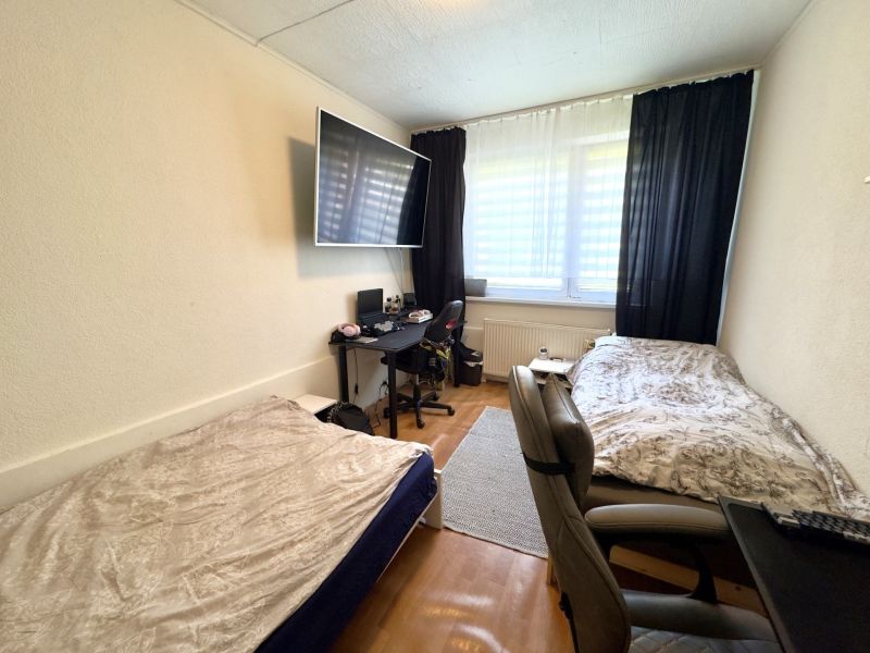 A room in a 4-room apartment with two beds, a wooden decor floor, and a work desk.