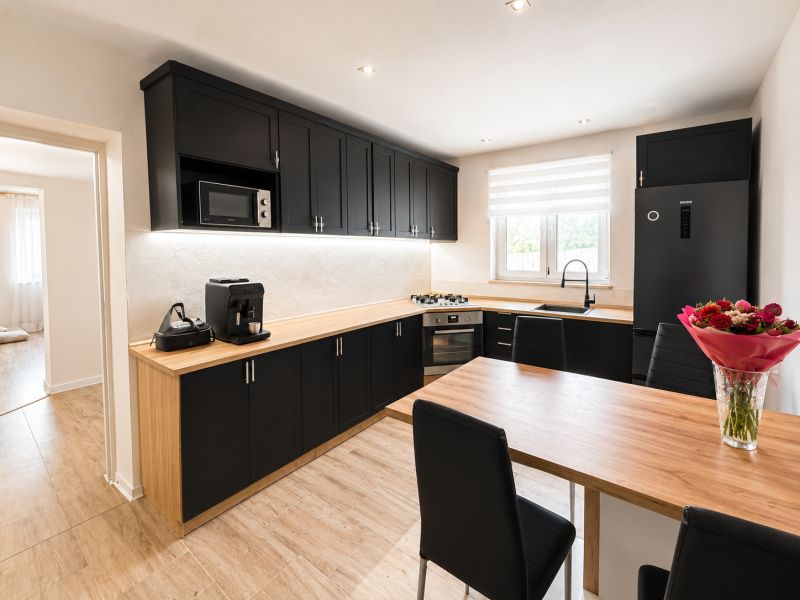 A kitchen in a family house with black cabinets, wooden floor decor, and a large window.
