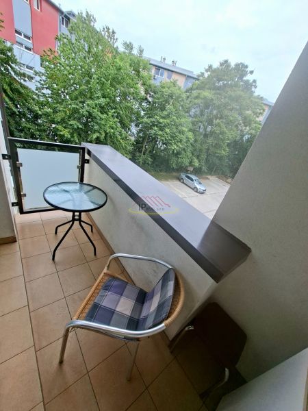 A studio apartment balcony with a view of greenery and a parking lot, a room with a table and a chair.