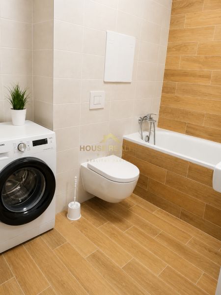 Bathroom with a toilet, bathtub, washing machine, and wooden decor flooring in a studio apartment.