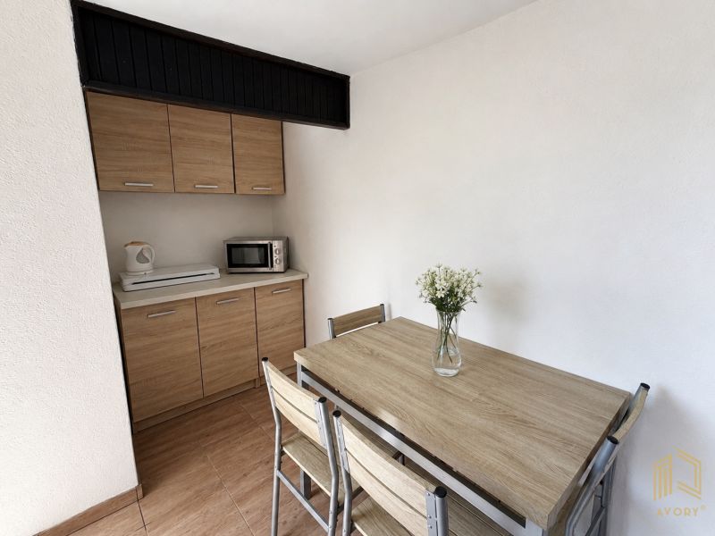 A kitchen in a cottage with a wood-patterned floor, a kitchen unit, and a table with a flower.
