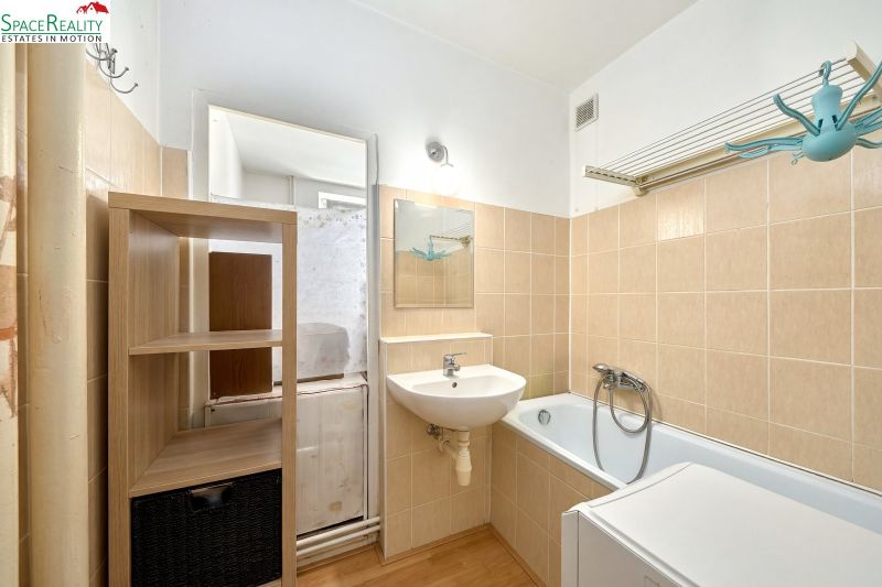 A bathroom with a bathtub and a sink in a 2-room apartment, light tiles, shelves.
