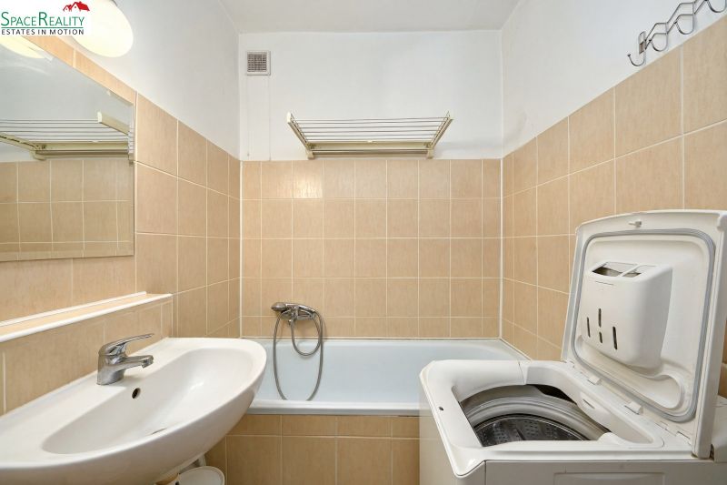 A bathroom with a washing machine and a bathtub in a 2-room apartment with beige tiles.