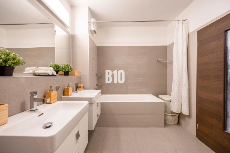 A bathroom in a 3-room apartment with a bathtub, two sinks, and neutral tiles.
