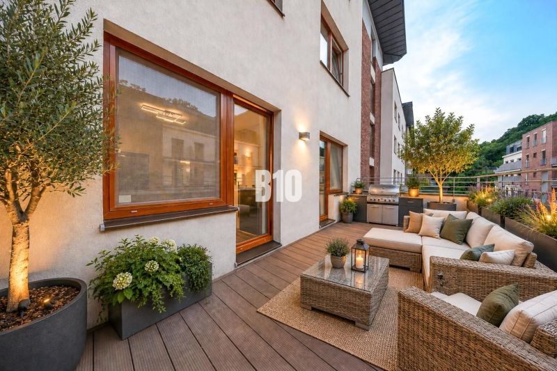 Terrace of a 3-room apartment with seating, plants, and a grill, floor with wooden decor.