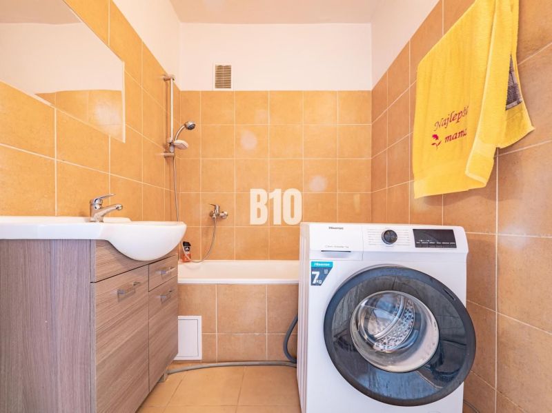 A bathroom in a 3-room apartment with a washing machine, a bathtub, and light tiles.