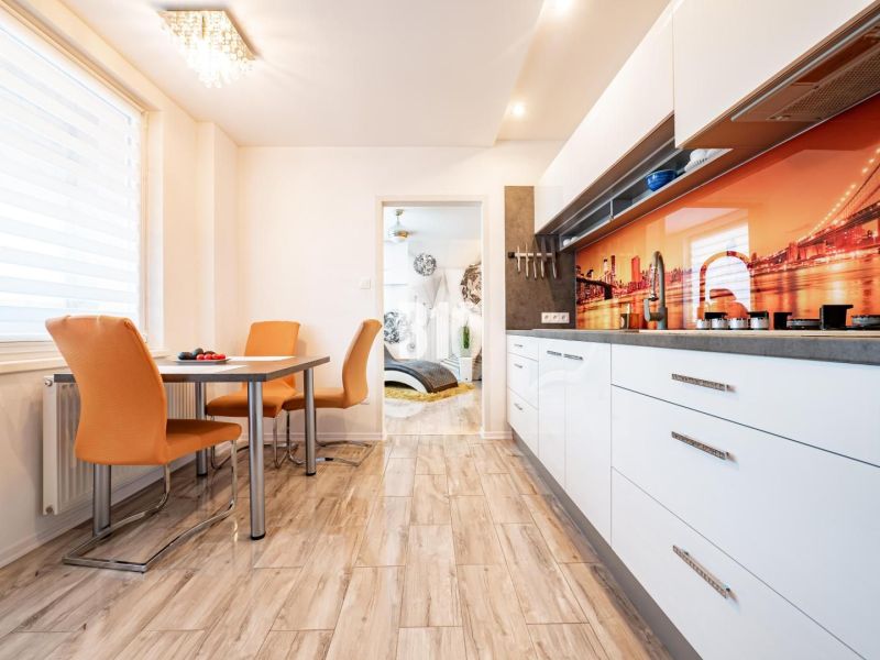 A kitchen in a 3-room apartment with orange chairs, a wooden decor floor, and colorful tiles.