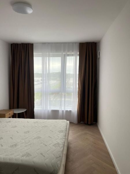 A bedroom in a 2-room apartment with a wood-patterned floor, simple furniture, and a large window.