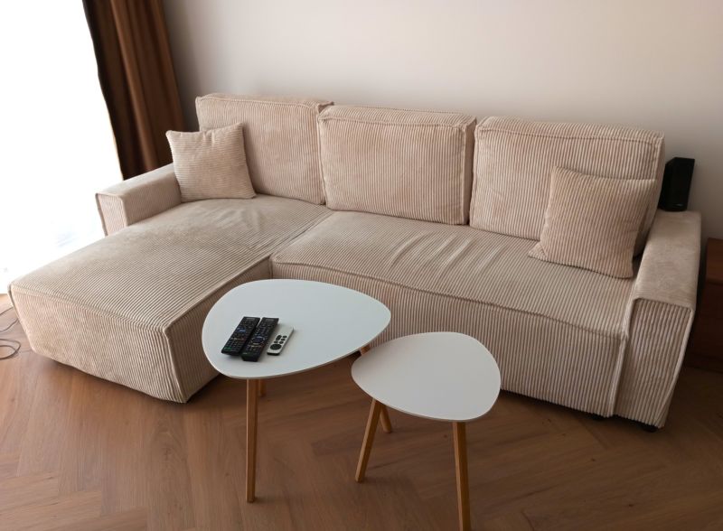 A light sofa with cushions, a pair of tables, a floor with a wooden decor, remote controls.
