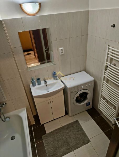 A bathroom in a studio apartment with a washing machine, sink, bathtub, and light-colored tiles.