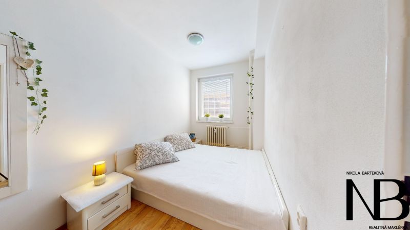 Bedroom in a 2-room apartment with a white bed, nightstand, and wooden decor flooring.