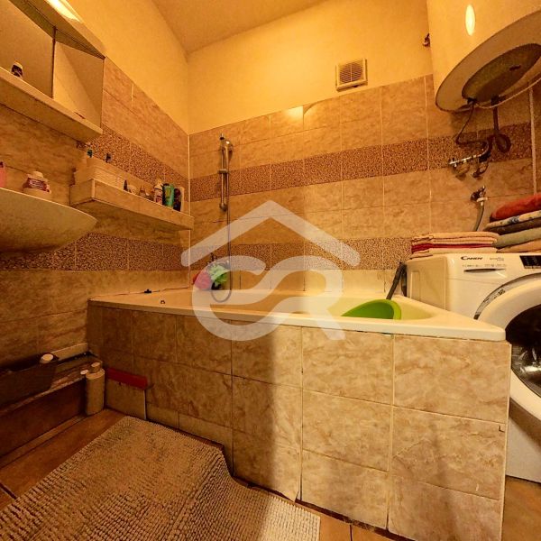 A bathroom in a family house with a bathtub, a washing machine, shelves, and light tiles.