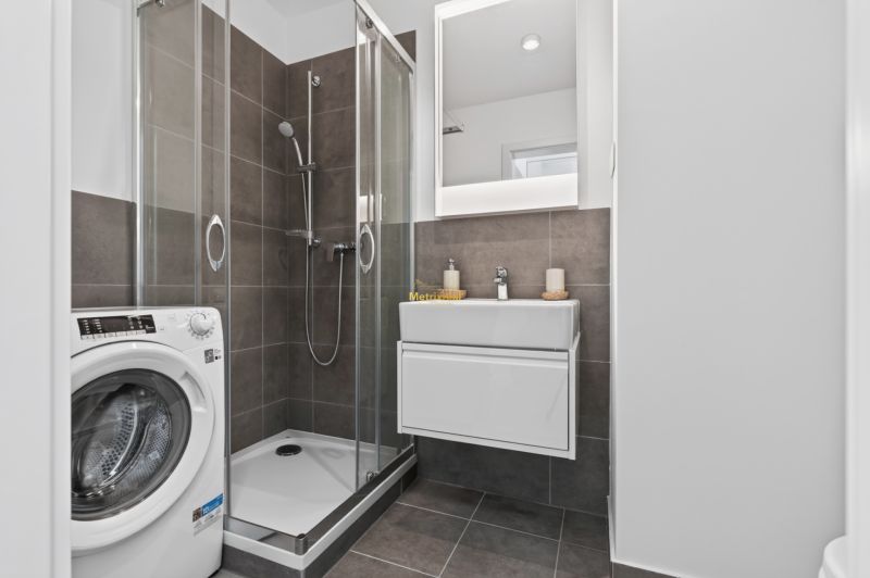A bathroom in a studio apartment with a washing machine, shower, and sink with a lighted mirror.