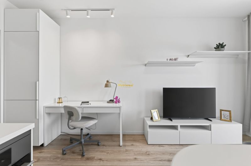 Studio apartment with white furniture, a desk, a television, and a floor with a wooden decor.