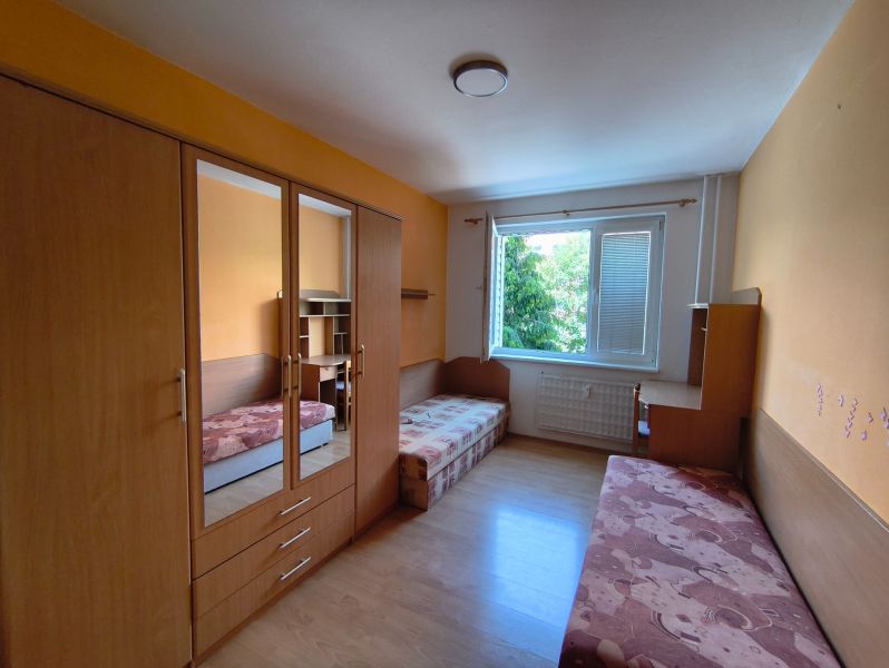 A room in a 3-room apartment with a wooden decor floor, a large window, and furniture.