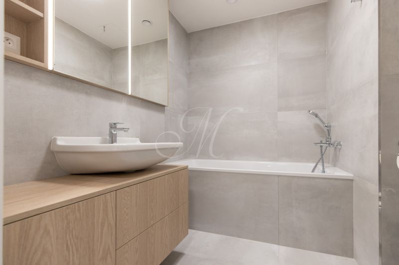 Bathroom with a bathtub, a sink on a wooden cabinet, a mirror, and light tiles in a studio apartment.