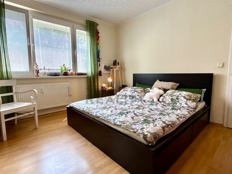 Bedroom in a 3-room apartment with a bed, wood-patterned flooring, and a white chair.