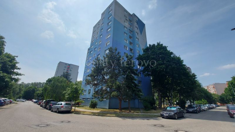 A taller residential apartment building in Bratislava - Petržalka surrounded by greenery and parked cars.