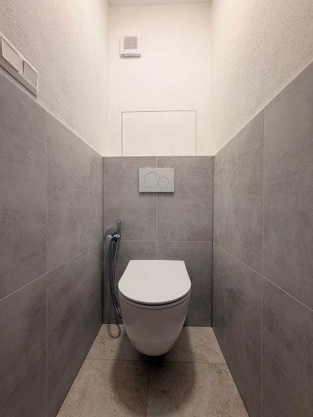 Bathroom in a 2-room apartment with a toilet, gray tiles, and a wall-mounted flush unit.
