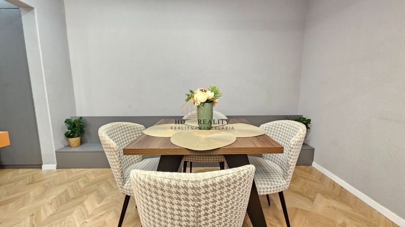 Dining area with flowers and chairs in a 2-room apartment with wood-patterned flooring.