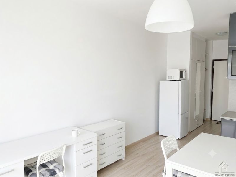 A studio apartment with wood-patterned flooring, white furniture, and a kitchen.
