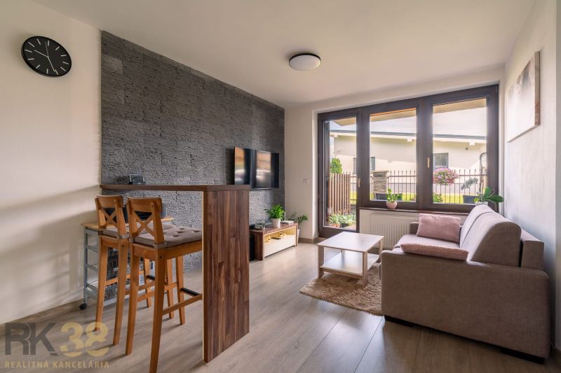 Living area of a 2-room apartment with a window, sofa, TV, and flooring with a wood decor.