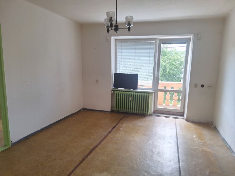 An empty room in a two-room apartment with a wood-patterned floor and a window to the balcony.