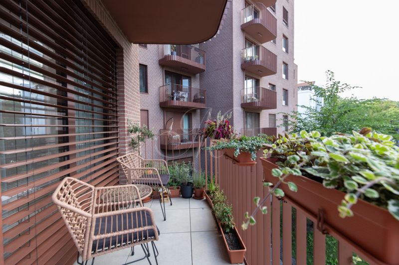 A balcony with chairs and flower pots in a 2-room apartment, overlooking a green area.