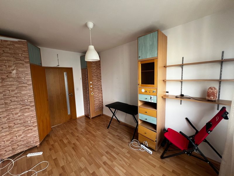 A room in a 3-room apartment with a wooden decor floor, shelves, and a relaxation chair.