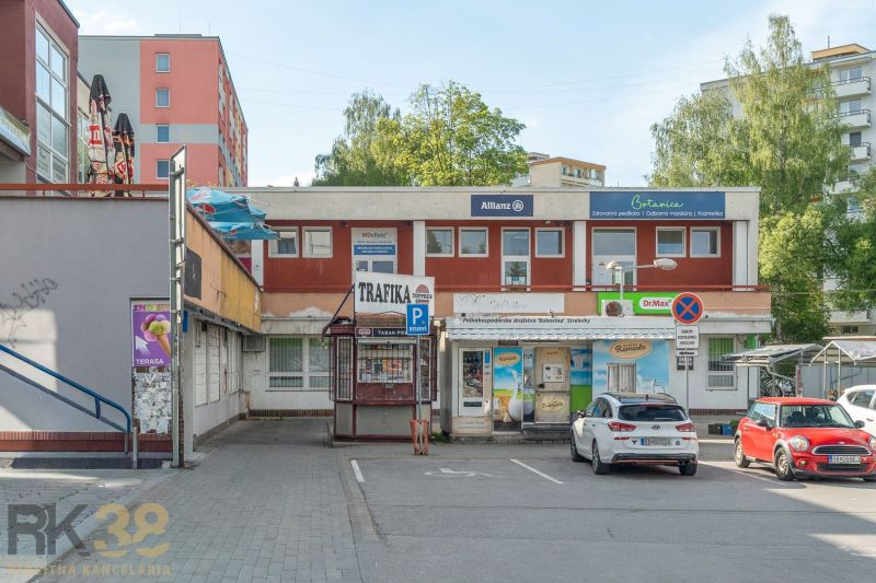 A building in Banská Bystrica with shops and a parking lot, surrounded by greenery and apartment blocks.