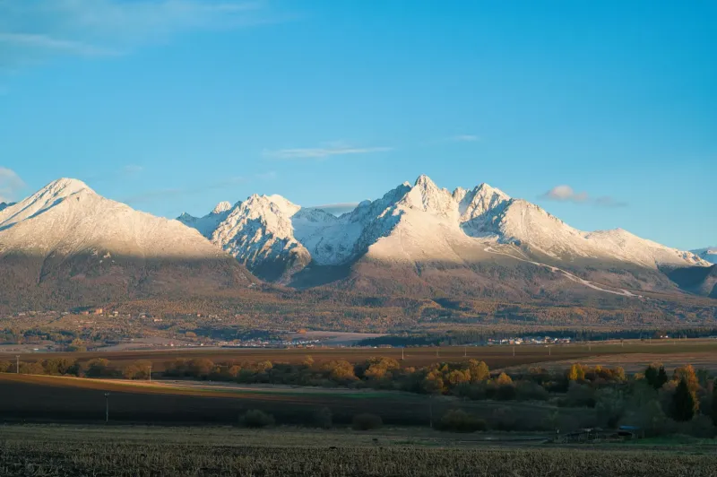 Poprad agricultural and forest lands with a panorama of snow-covered Tatra peaks under a blue sky.