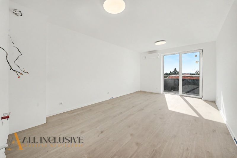 Bright living room in a two-room apartment with wood-patterned flooring and large windows.