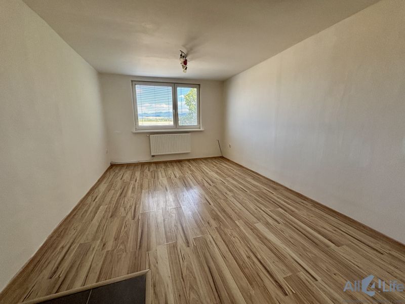 A room in a three-room apartment with a wood-patterned floor and a large window.