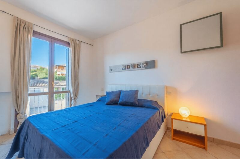 A bedroom in a holiday apartment with a double bed, a window, and a nightstand.