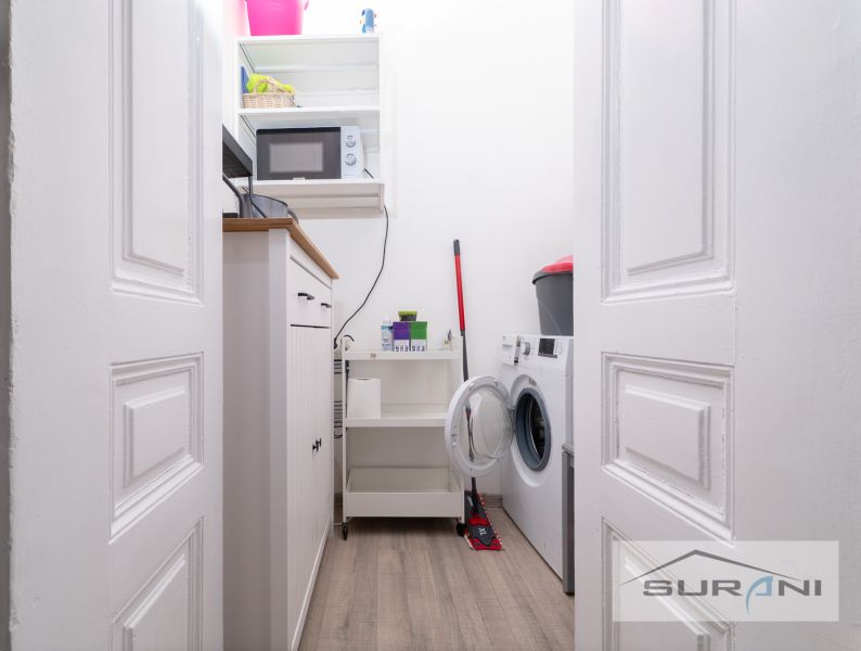 Laundry room in a 2-room apartment with a washing machine, shelf, microwave, and wooden decor flooring.