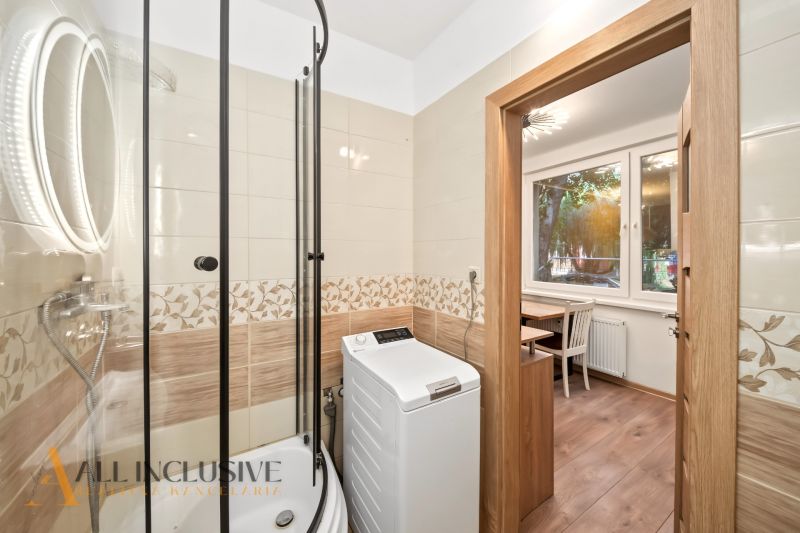 Bathroom with a washing machine and a shower in a 2-room apartment, with a view into the room with a wooden decor floor.