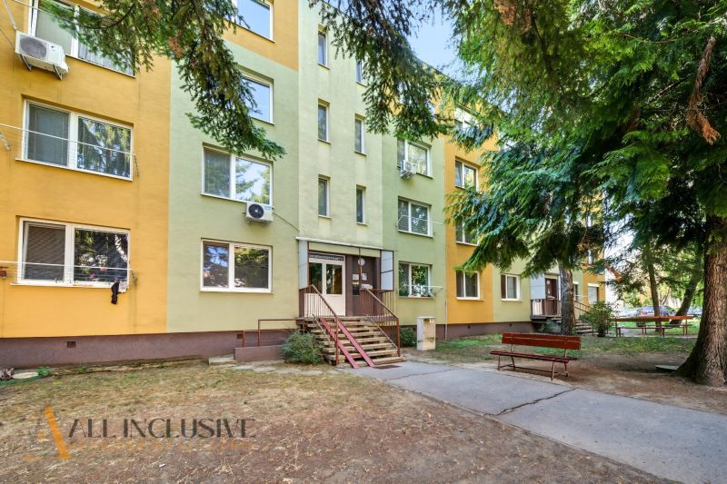 The entrance to the apartment building on Nešporova Street in Šaľa, with a bench and trees.