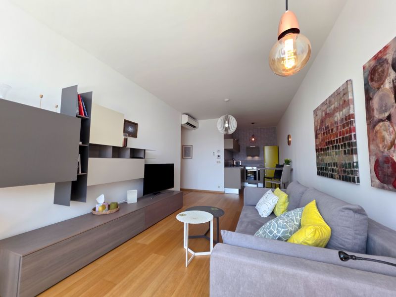 Living area of a studio apartment with a sofa, decorative cushions, and a wooden-patterned floor.