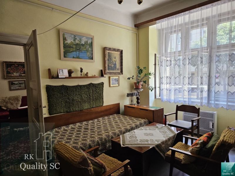 Living room of a two-room apartment with a sofa, paintings, and a large window with curtains.