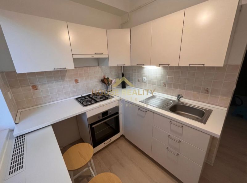 A kitchen with white cabinets, a built-in oven, and counter stools in a studio apartment.