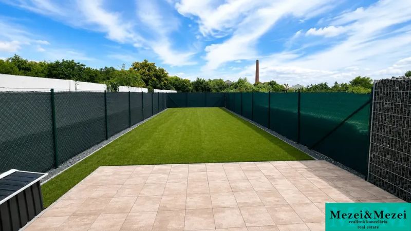 The garden of a family house in Trnava with a lawn and a paved terrace under a blue sky.